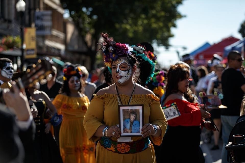 Tanya Castilleja Perez in a yellow off-shoulder dress with sugar skull face paint and a flower crown, walking in the festival parade holding a framed photo close to her heart